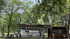 Een rustiek RV-park met een overdekte picknickplaats, picknicktafels en een klein gebouw met een veranda.