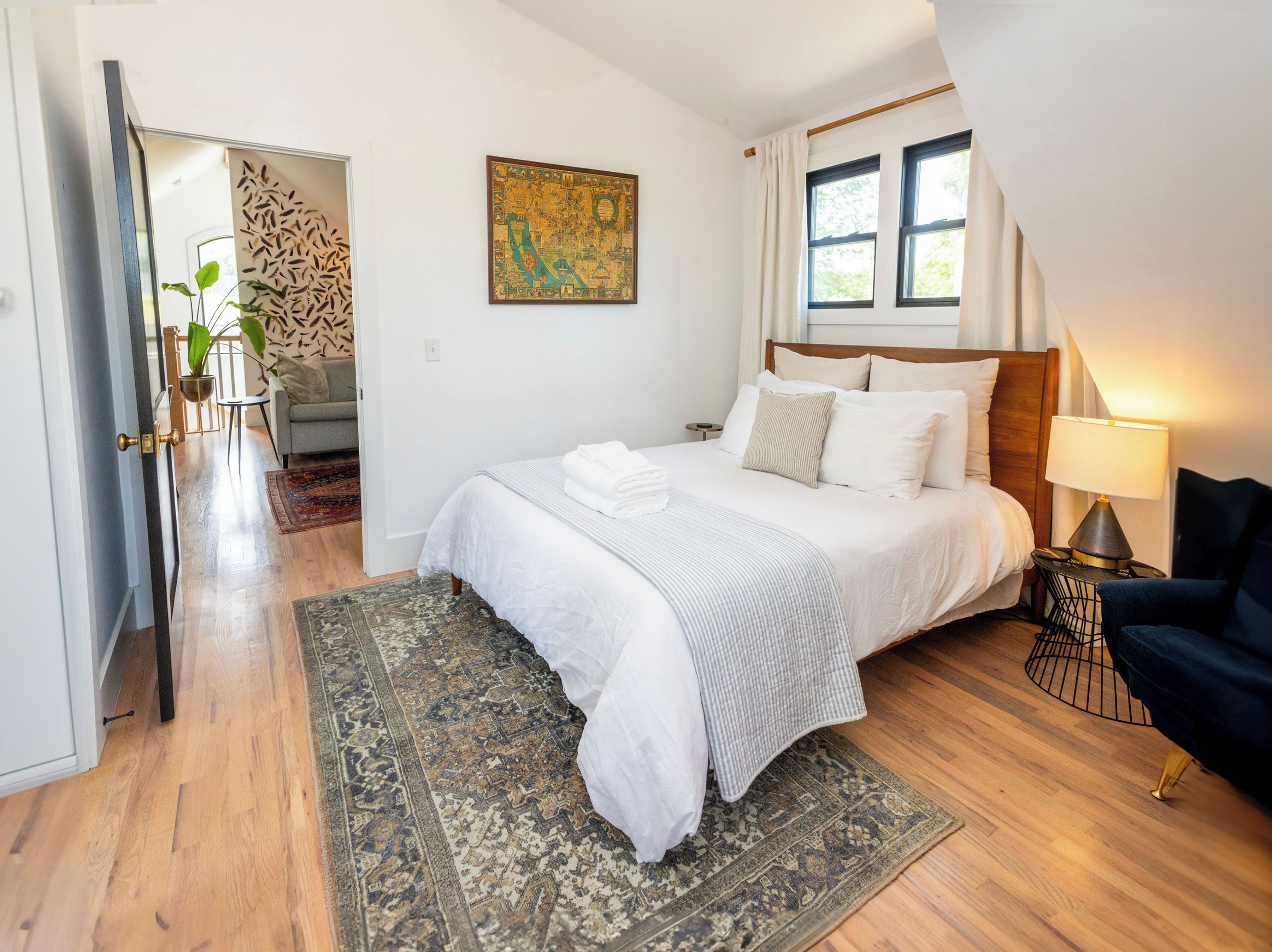 Un dormitorio con una cama, una mesita de noche, una lámpara, una silla, un cuadro y una puerta que da a otra habitación.