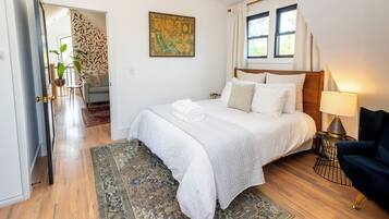 A bedroom with a bed, a nightstand, a lamp, a chair, a painting, and a door leading to another room.