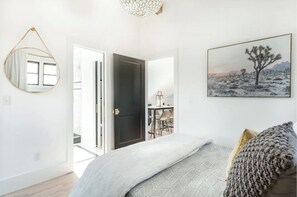 A bedroom with a bed, a round mirror, a door leading to a dining area, and a painting of a desert landscape.