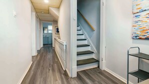 Un couloir avec un plancher en bois, des murs blancs, un escalier aux marches sombres et une étagère en métal sur le côté droit.
