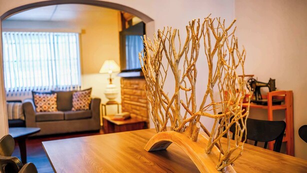 Une table à manger avec un élément décoratif en bois au centre, un canapé, une lampe et un téléviseur à l’arrière-plan.