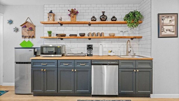 Een moderne keuken met grijze kastjes, een roestvrijstalen vaatwasser, een magnetron, en houten planken met decoratieve objecten.