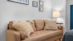 Een beige bank met gekleurde kussens, een lijst met foto's aan de muur en een tafellamp.
