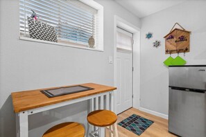 A compact kitchen with a wooden counter, two stools, a refrigerator, a window with blinds, and a decorative hanging on the wall.