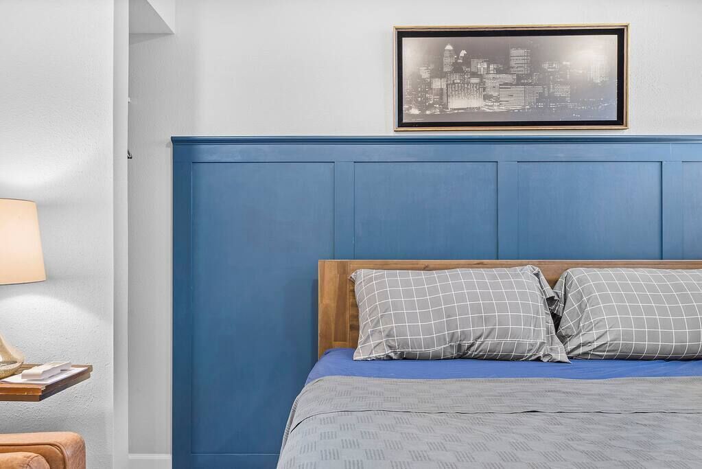 A bedroom with a blue headboard, a bed with grey checkered bedding, a framed cityscape picture on the wall, and a bedside table with a lamp and a small item on it.