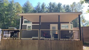 A two-story building with a covered patio area, a fenced-in section with a grill, and a view of trees in the background. - The Honey Crisp @ Backyard Apple Cottages (FLAT ROCK)