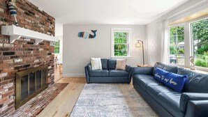 Een woonkamer met een haard van baksteen, een blauwe leren bank en uitzicht op groen.