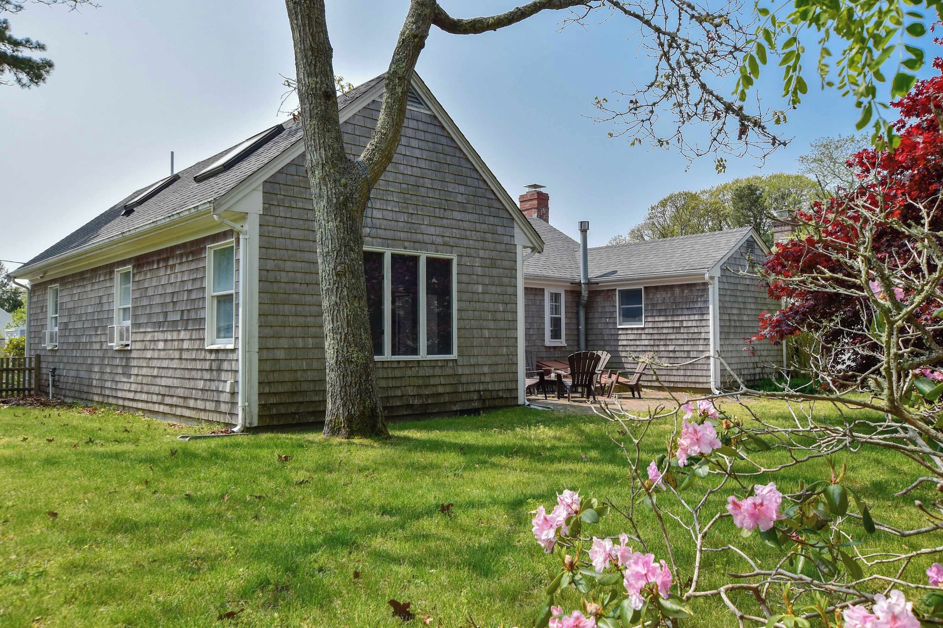 A two-story house with a sloped roof, a visible chimney, and a front yard with a lawn and flowering shrubbery.