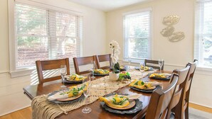 A dining room with a wooden table set for a meal, surrounded by chairs, with windows and decorative wall art. - Spacious Stone Fire Pit Game Room! (Marstons Mills)