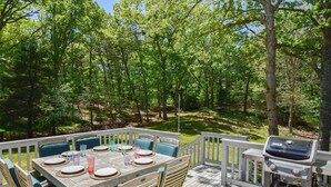 A wooden deck with a table set for four, surrounded by chairs, and a barbecue grill. - Spacious Stone Fire Pit Game Room! (Marstons Mills)