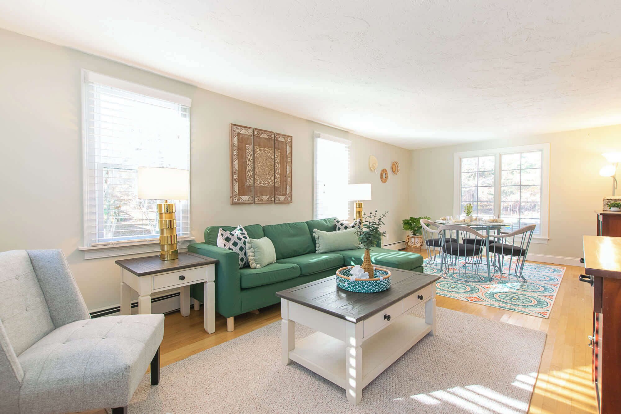 A living room with a green sofa, a white coffee table, a grey armchair, a dining table with chairs, and a window with blinds.