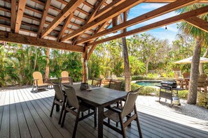 A covered outdoor area with a wooden deck, a dining table with chairs, a grill, and a pool in the background. - Shady Mango by Duncan Real Estate (Anna Maria)