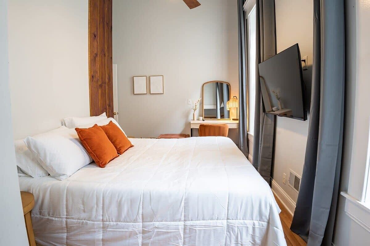 Un lit bien fait, agrémenté d’oreillers blancs et orange, une table de chevet avec une lampe, un miroir et un téléviseur fixé au mur.