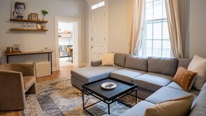 Una sala de estar con un sofá modular, una mesa de centro y una alfombra. Se ve una puerta que da a otra habitación y una ventana con cortinas.