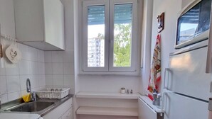 Una cocina compacta con armarios blancos, horno integrado, lavamanos y ventana con persianas.