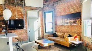 A modern living space with a brick wall, a sofa, a coffee table, and a TV mounted on the wall. - Historic Penthouse Style Loft in OTR, Parking Included (Cincinnati)
