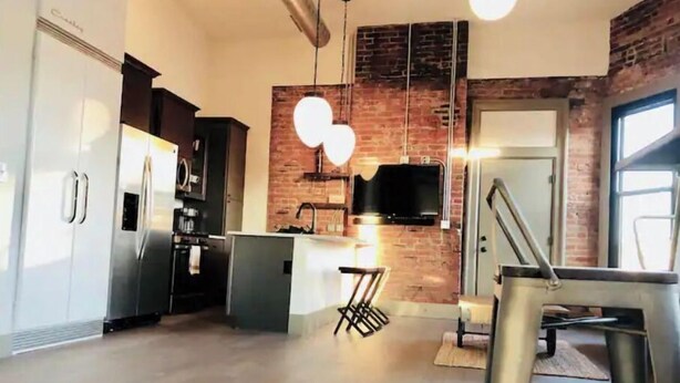 A modern kitchen with a brick accent wall, stainless steel appliances, and a flat-screen TV mounted above the island. - Historic Penthouse Style Loft in OTR, Parking Included (Cincinnati)
