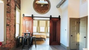 A room with a loft bed, a dining area with a table and chairs, a brick wall, and a large window. - Historic Penthouse Style Loft in OTR, Parking Included (Cincinnati)
