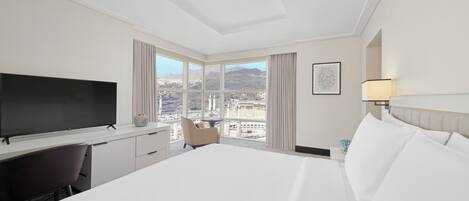 A hotel room with a large bed, a desk with a flat-screen TV, a chair, a window with a city view, and a lamp.