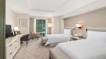 A hotel room with two beds, a desk with a TV, a chair, a small table, and a window with curtains.