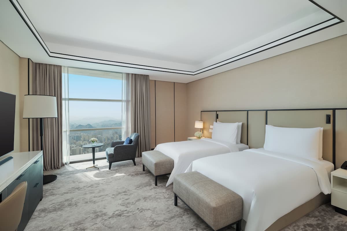A hotel room with two beds, a desk, a chair, a TV, and a view of a cityscape.