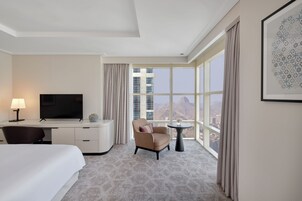 Habitación de hotel con cama, escritorio con televisor, silla, mesita pequeña y ventana grande que ofrece vistas a la ciudad.