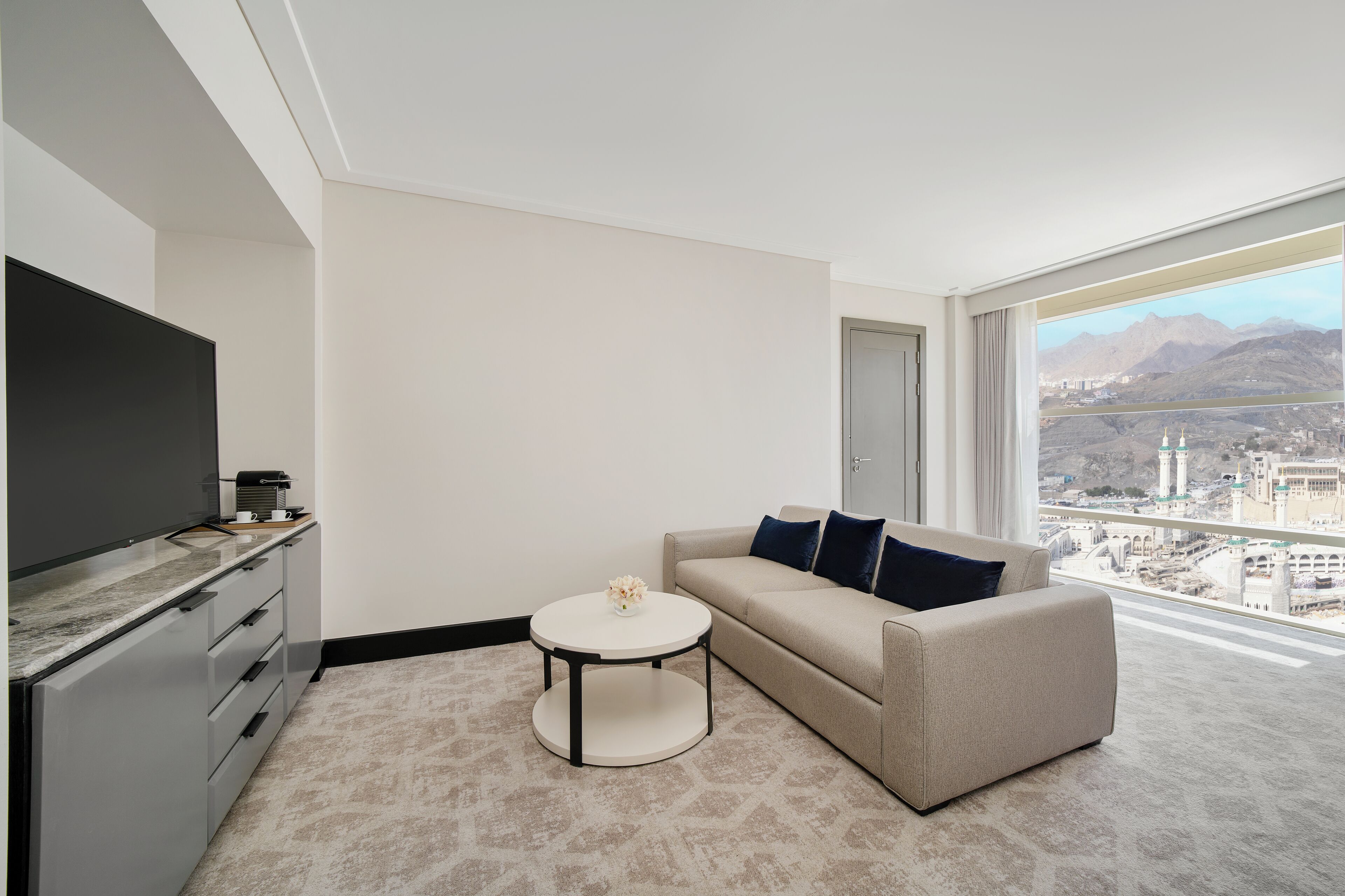 A modern hotel room with a sofa, a coffee table, a flat-screen TV, and a large window offering a city view.