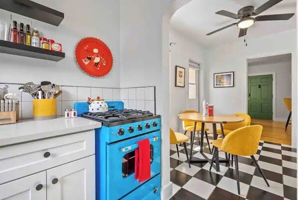 A kitchen with a blue stove, white cabinets, and a red wall decoration. There is a dining area with yellow chairs and a checkered floor.