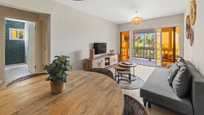 A spacious living area with a wooden dining table, a grey sofa, wicker chairs, and a television on a wooden cabinet.