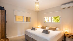 A bedroom with a large bed, two bedside tables, a hanging light fixture, and a window with a view of trees.