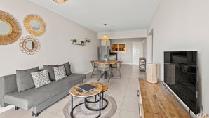 A modern living room with a grey sofa, round coffee table, and a TV. There is a dining area with a table and chairs, and a kitchen in the background.