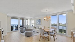 A spacious living room with a dining area, large windows, and a balcony with a view. - Beautiful 3 bedroom beachfront corner unit-CL0804 (Naples)