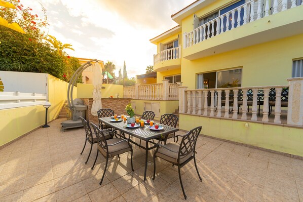 A dining area with a table set for six, surrounded by chairs, on a tiled patio with a yellow building in the background.