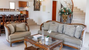 A spacious living room with a sofa, armchair, and coffee table, a staircase, and a kitchen in the background.