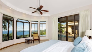Une chambre spacieuse avec un grand lit, un ventilateur de plafond et une vue sur une piscine et un paysage verdoyant, grâce à de grandes fenêtres.