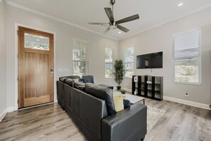 Un salon moderne avec un canapĂ© dâangle, un ventilateur de plafond, un tĂ©lĂ©viseur Ă Ă©cran plat et de grandes fenĂȘtres Ă©quipĂ©es de stores.