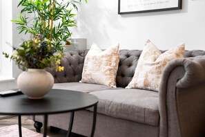 Un salón con un sofá gris, tres cojines, una mesa redonda con un florero y un cuadro en la pared.