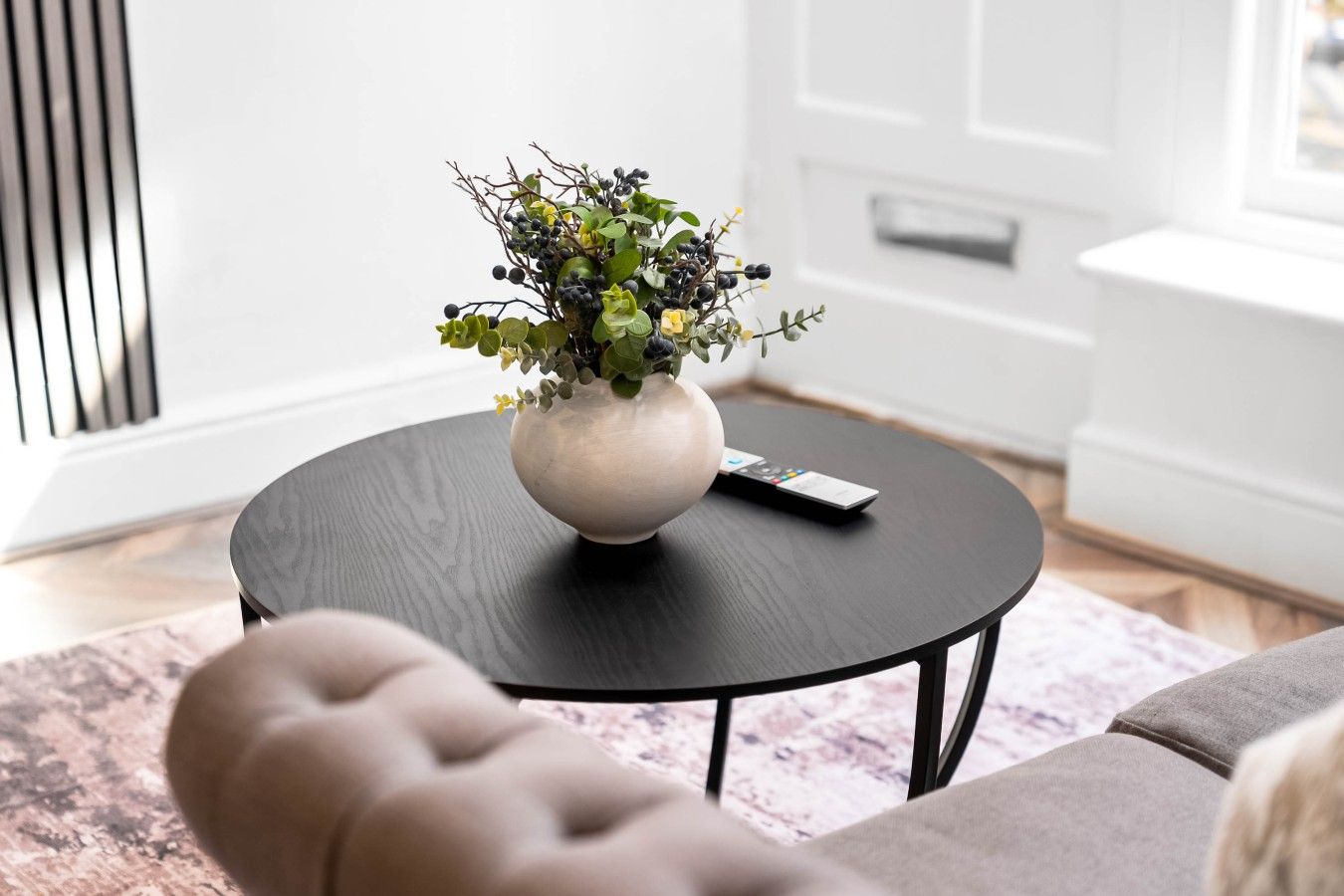 A living room with a round black coffee table, a beige sofa, and a white vase with green foliage and berries.