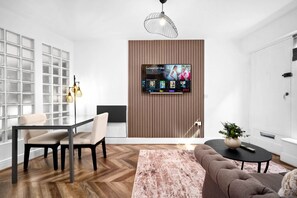 Ein modernes Wohnzimmer mit Flachbildfernseher an einer Wand aus Holzpaneelen, einem Essbereich mit Glastrennwand und einer Sitzecke mit Sofa und Couchtisch.