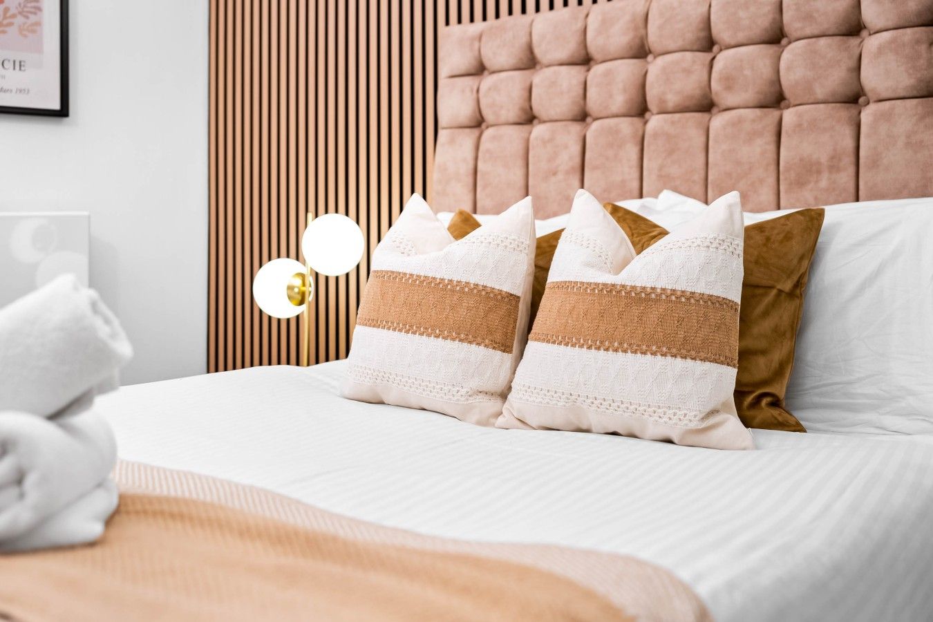 Cama bem arrumada com travesseiros brancos e marrons, cabeceira de madeira e luminária fixada na parede.