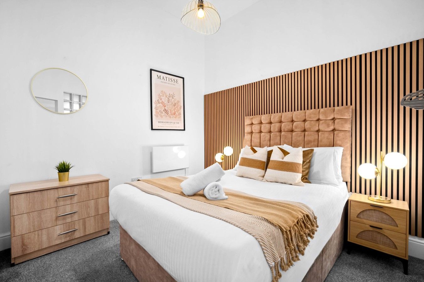 A modern bedroom with a tufted headboard, a bedside table with a lamp, a bedside plant, and a framed artwork on the wall.