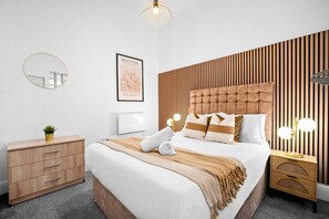 Un dormitorio moderno con un cabecero capitonado, una mesita de noche con lámpara, una planta junto a la cama y una obra de arte enmarcada en la pared.