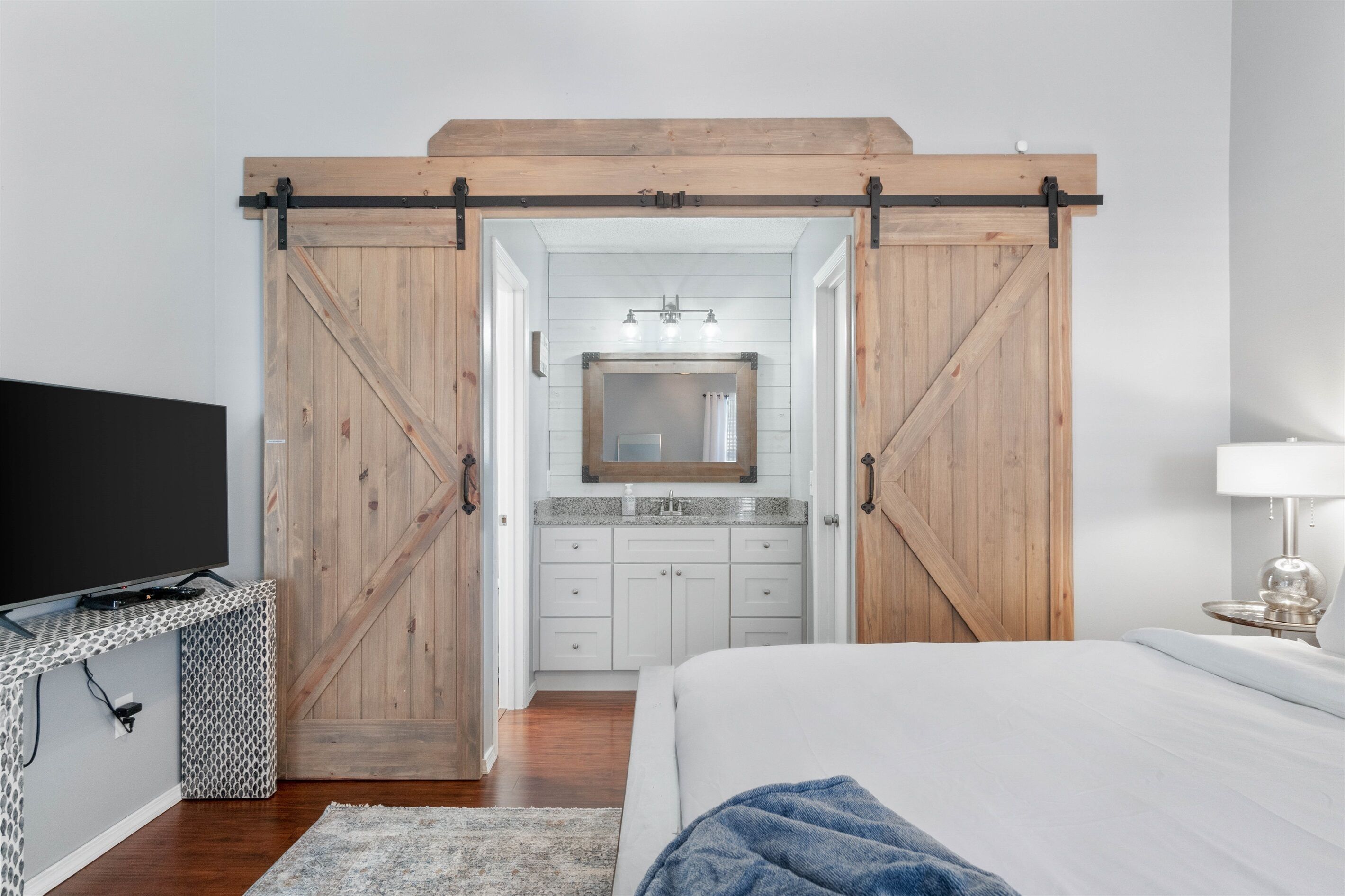 Quarto com uma cama grande, uma televisão sobre uma mesinha de cabeceira, banheiro com espelho e pia, e uma porta de correr estilo celeiro.