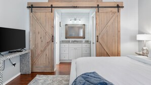 Un dormitorio con una cama grande, un televisor sobre una mesita de noche, un baño con espejo y lavamanos, y una puerta corredera de estilo rústico.