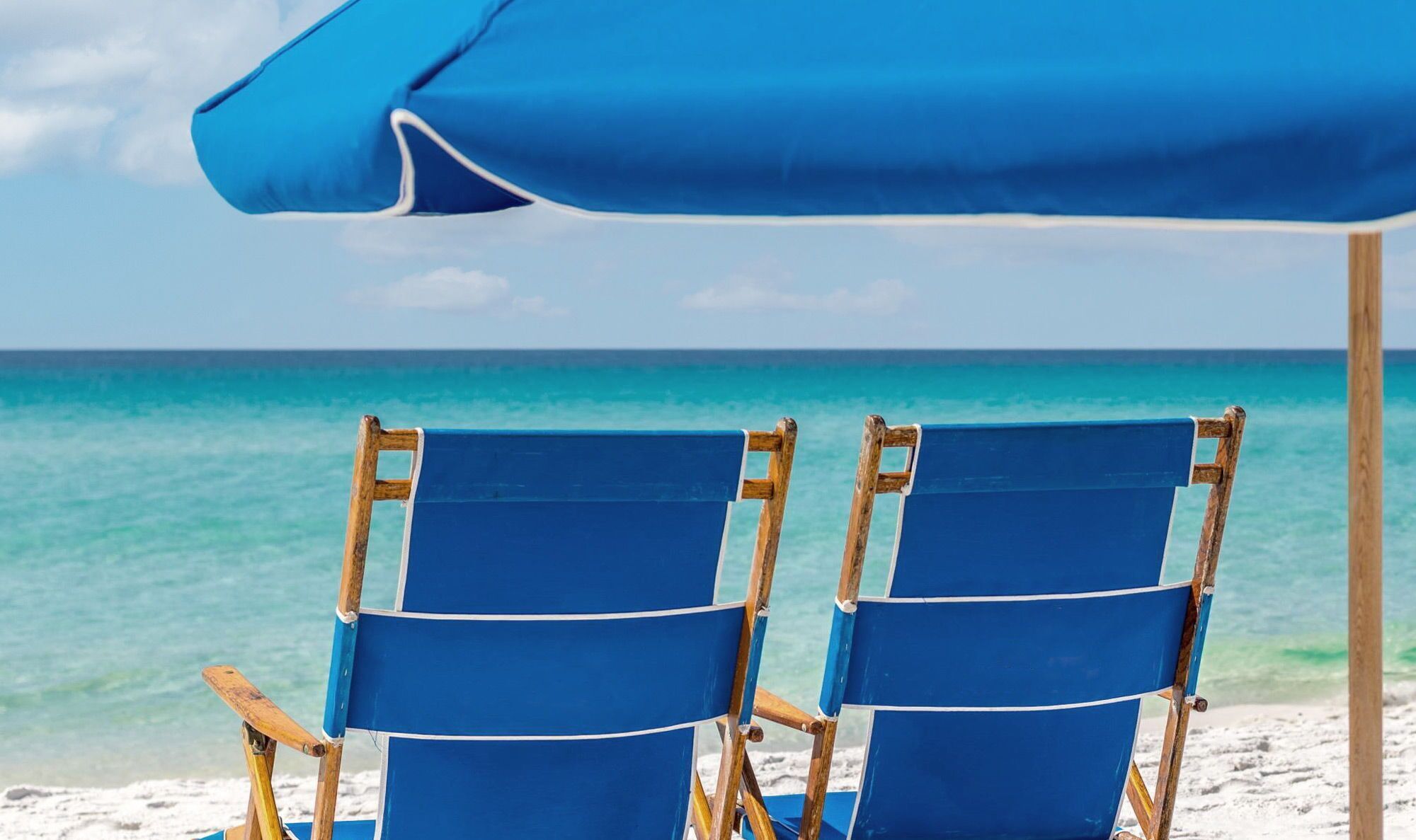 Deux chaises de plage bleues, placées sous un parasol bleu, sur une plage de sable fin bordée d’une eau turquoise et limpide.