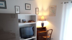 Ein Wohnzimmer mit einer Couch, einem Fernseher auf einem Regal, einer Lampe und einem Esstisch.