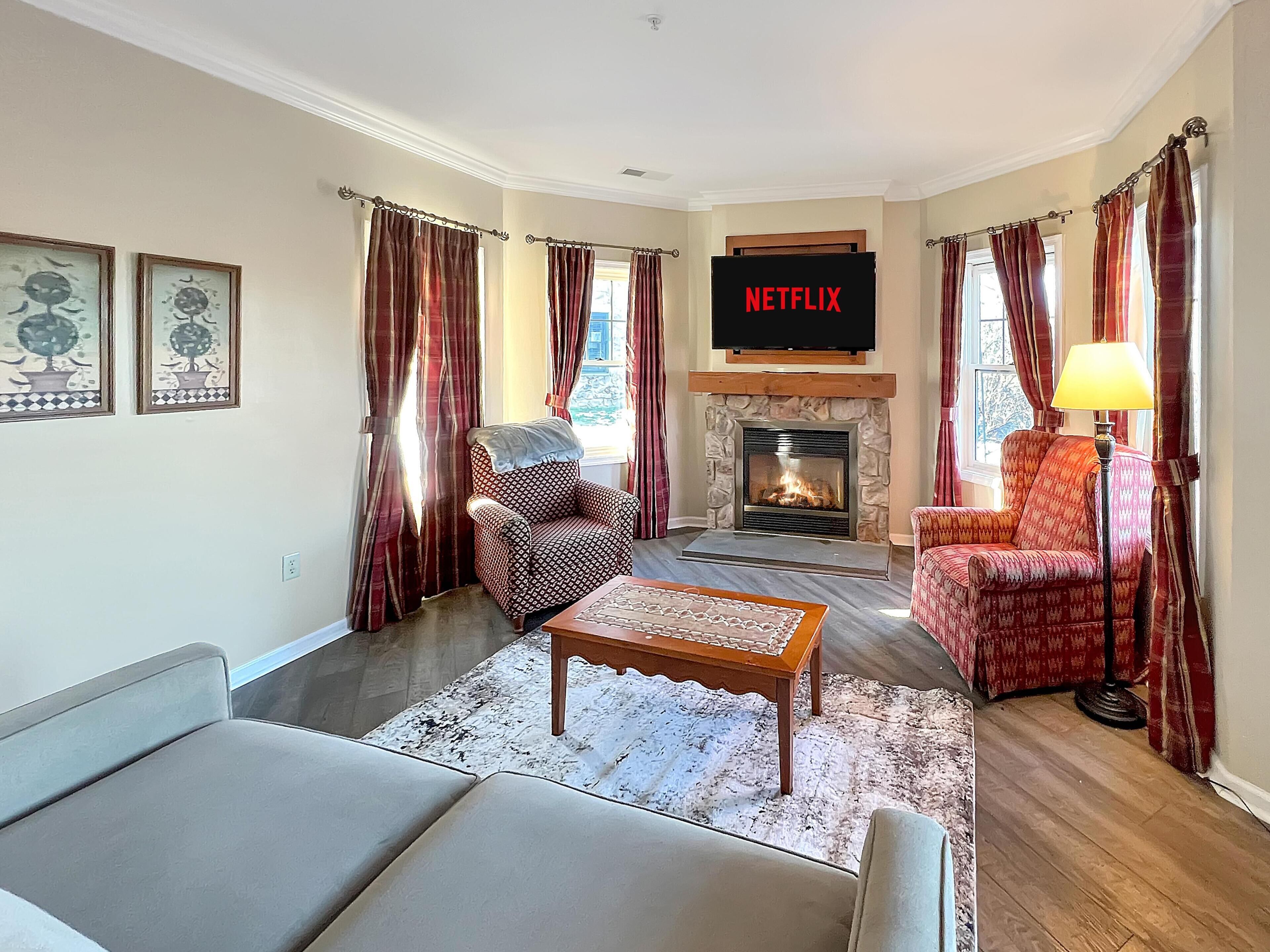 A living room with a fireplace, a sofa, a patterned armchair, a coffee table, and a television mounted above the fireplace.