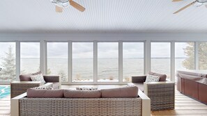 A spacious room with a large window overlooking a body of water, a ceiling fan, and a wicker sofa with cushions. - Smooth Sailing on the Lake! Free EV charging! (Plattsburgh)