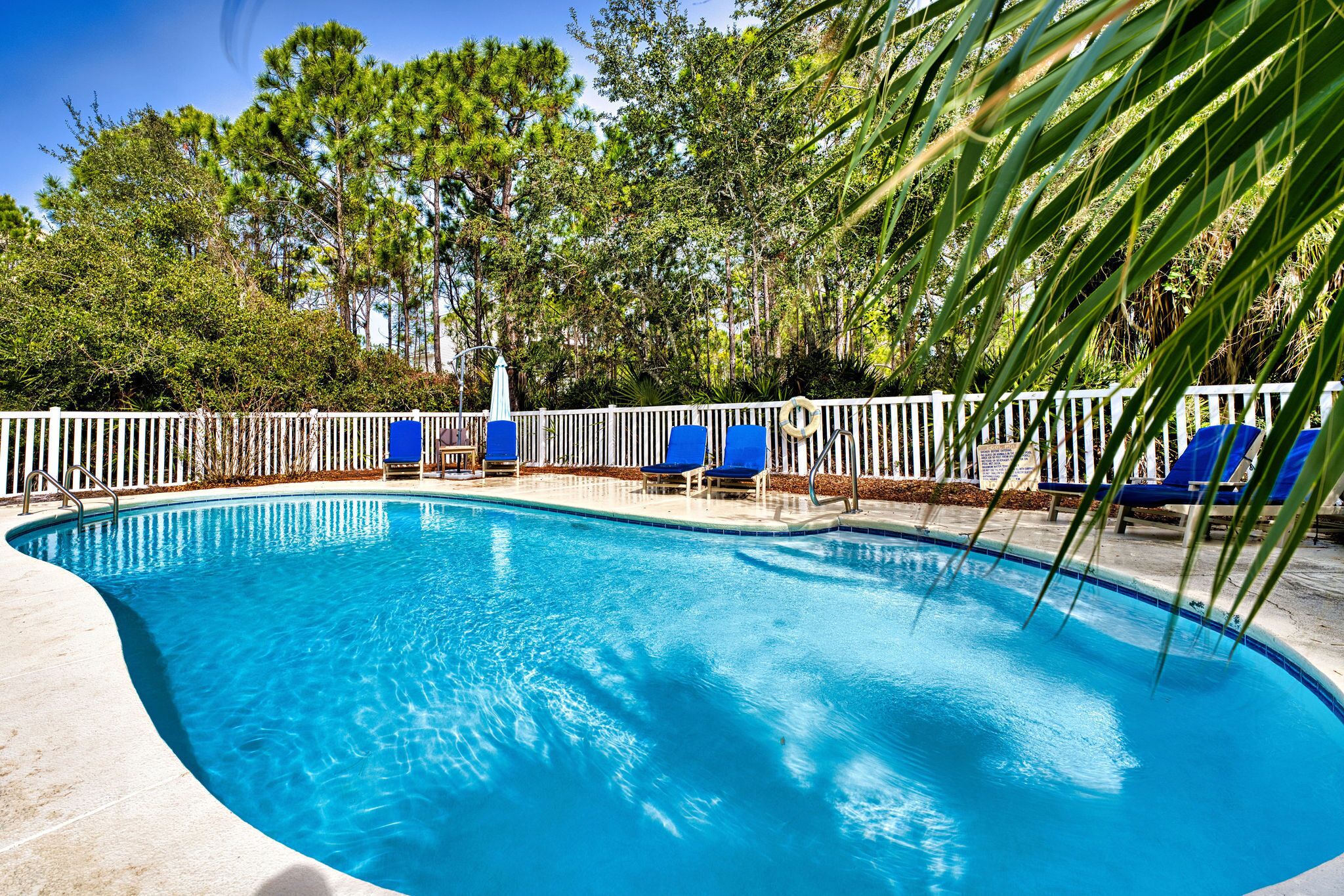 Une piscine à l’eau bleue, entourée d  ’une clôture blanche et de chaises longues bleues.
